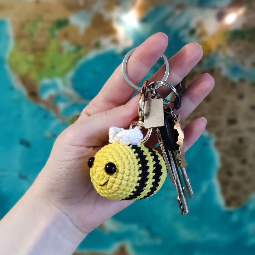 Hand holding a handmade crochet bee keychain amigurumi in front of a world map background.