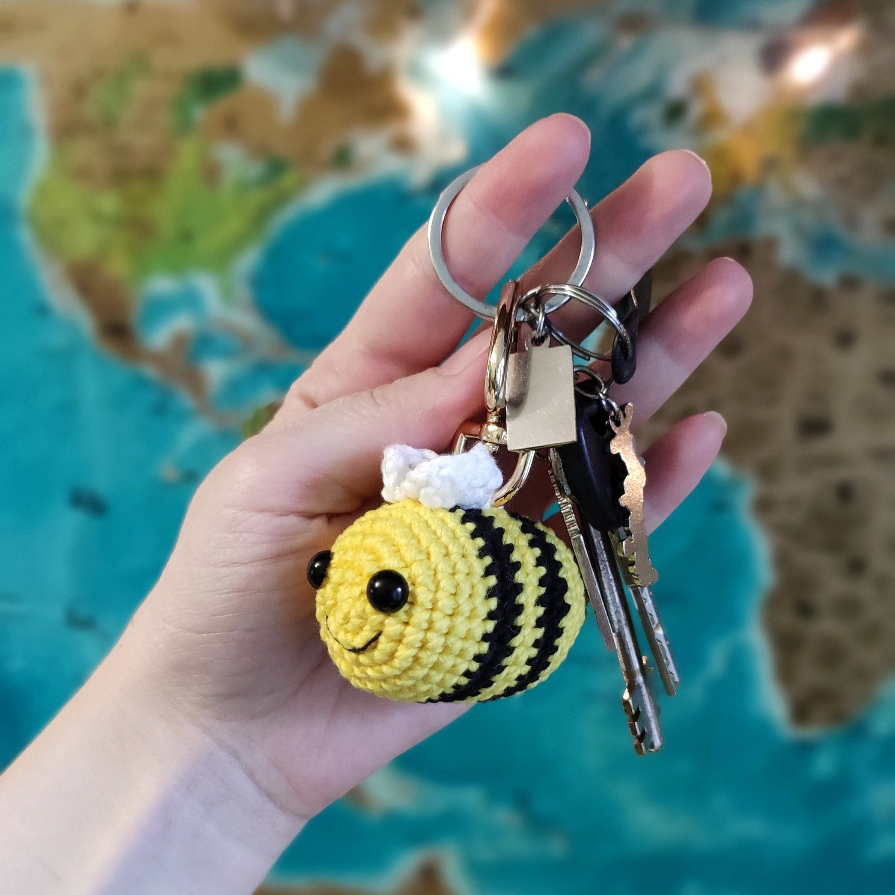 Hand holding a handmade crochet bee keychain amigurumi in front of a world map background.