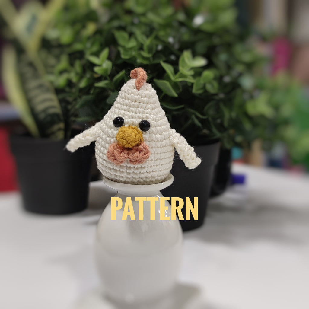 Handmade crochet chicken and rooster amigurumi sitting on a white stand with green plants in the background.