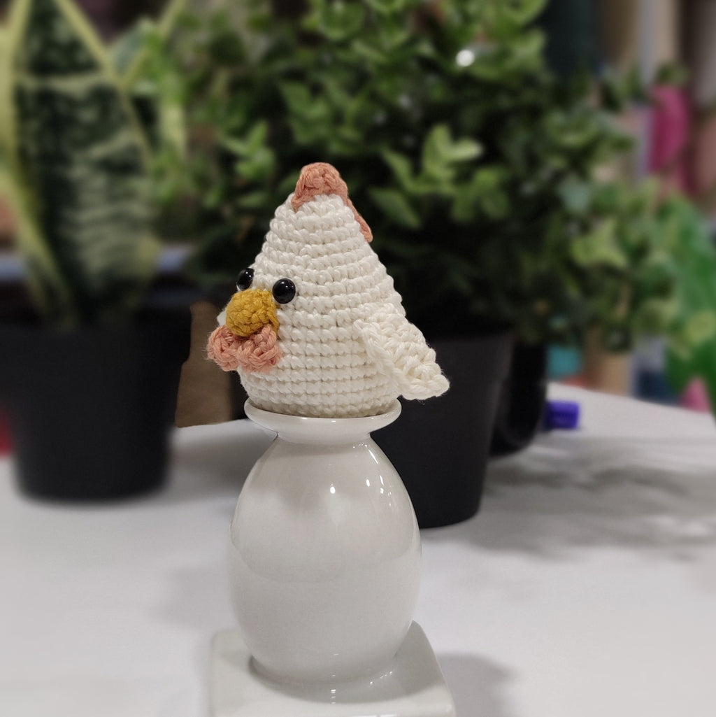 Handmade crochet chicken and rooster amigurumi sitting on a white stand with green plants in the background.