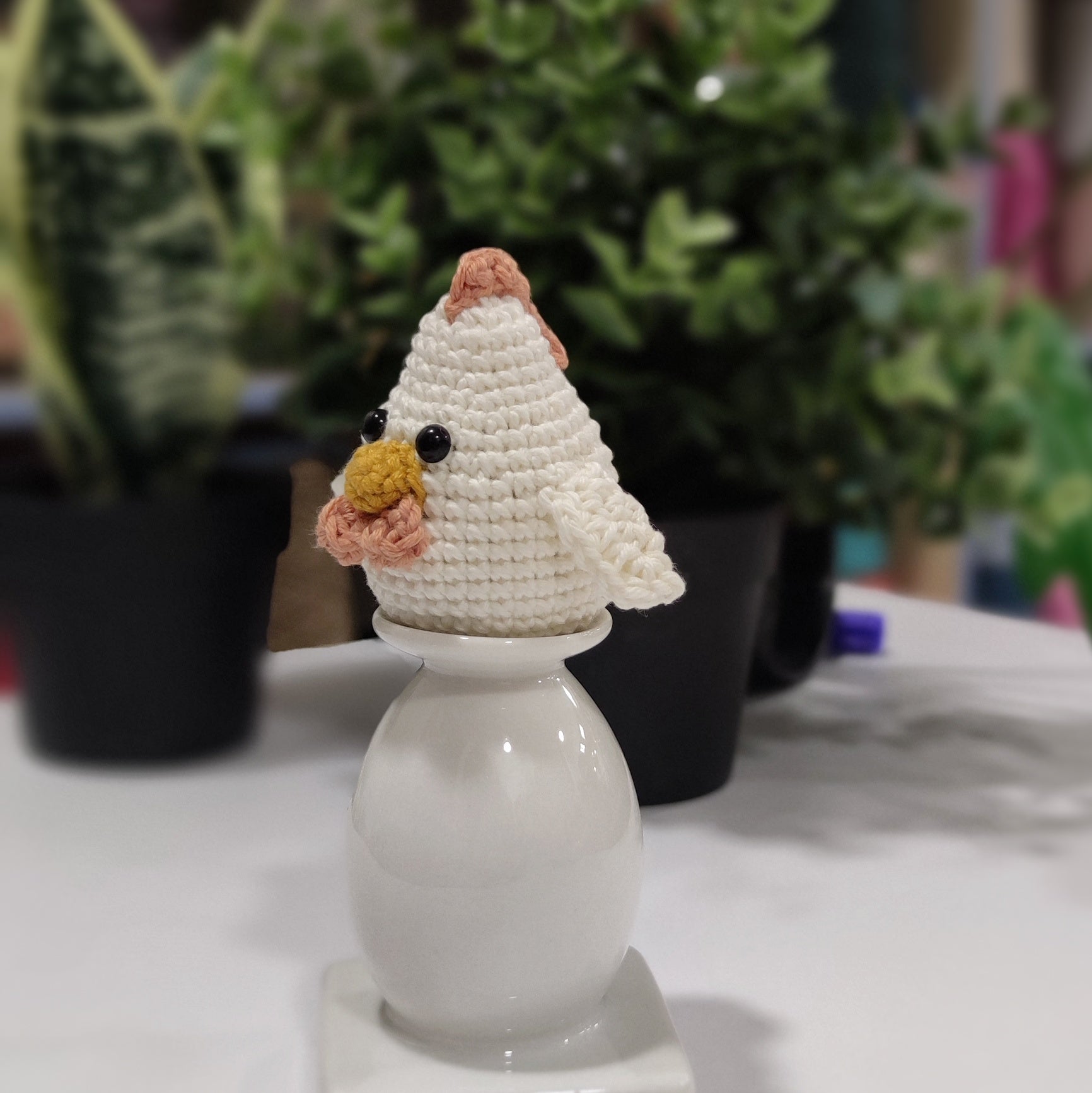 Handmade crochet chicken and rooster amigurumi sitting on a white stand with green plants in the background.