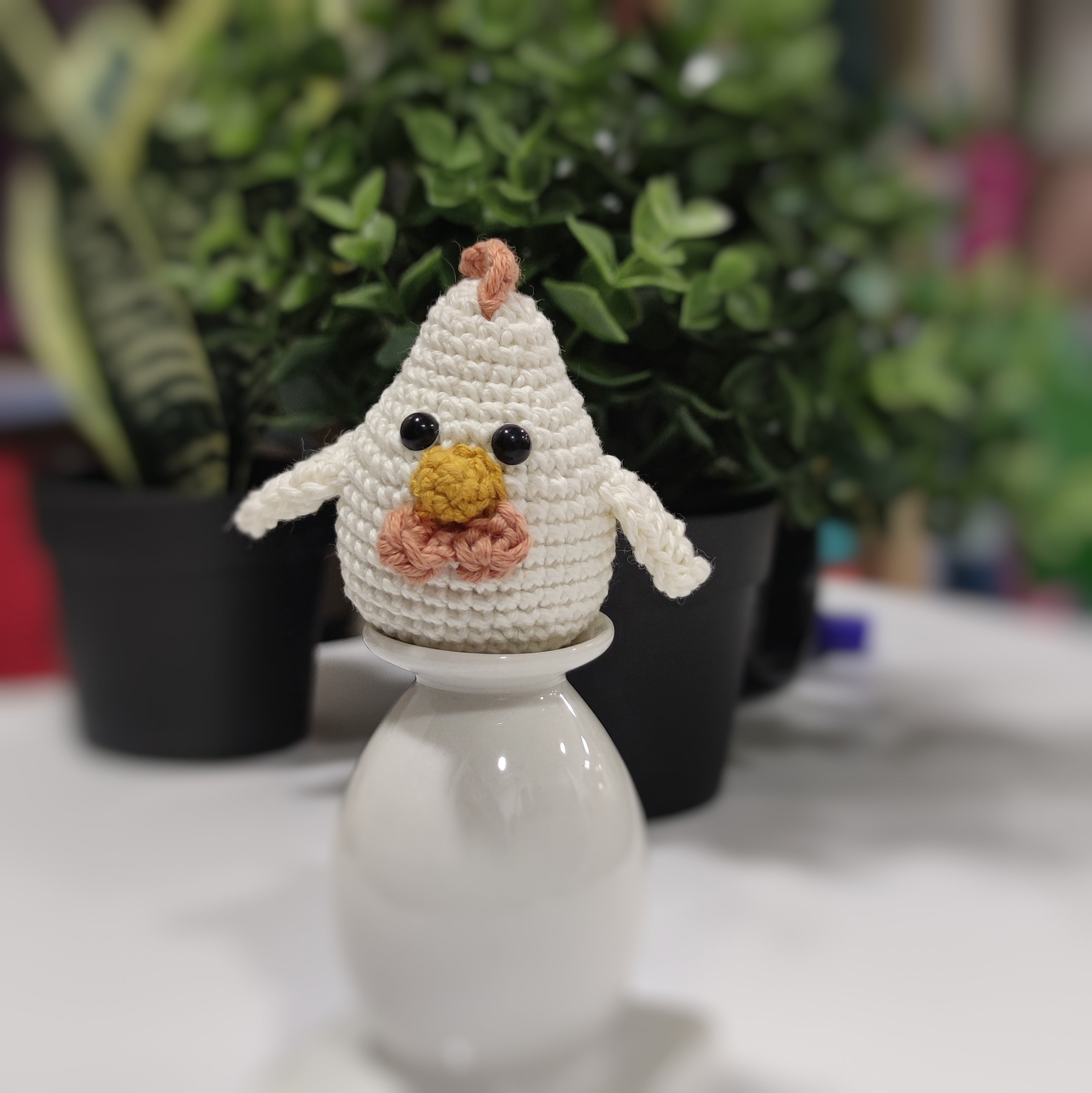 Handmade crochet chicken and rooster amigurumi sitting on a white stand with green plants in the background.