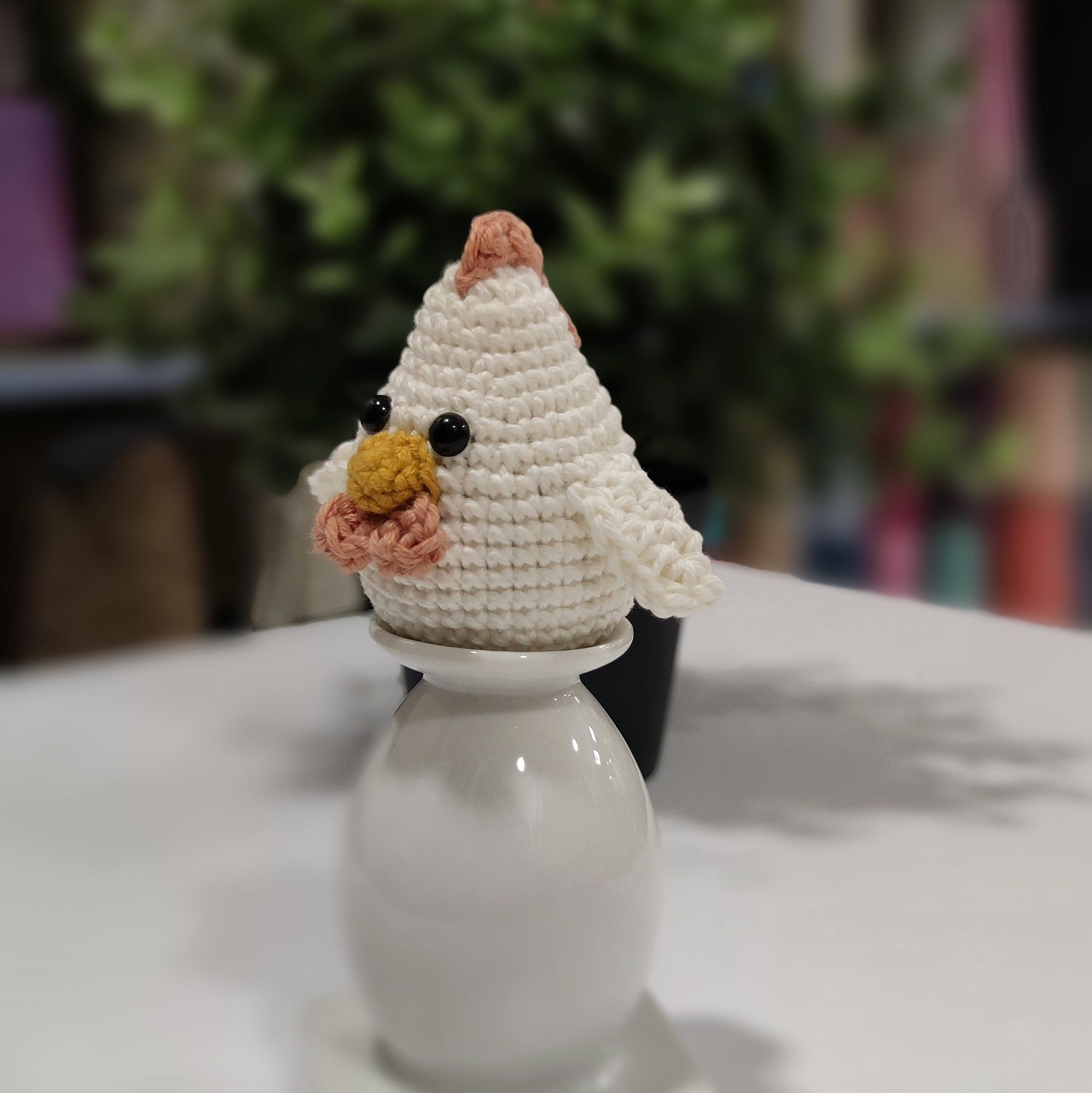 Handmade crochet chicken and rooster amigurumi sitting on a white stand with green plants in the background.
