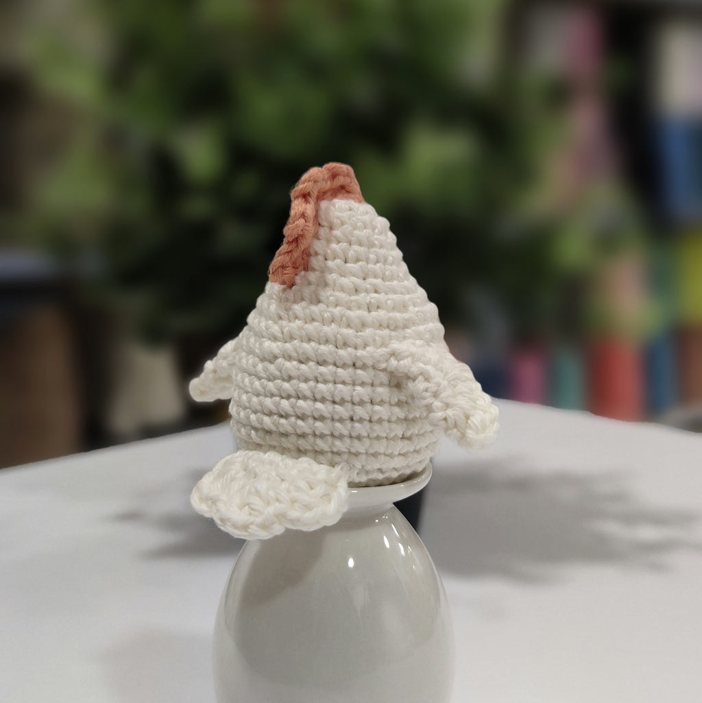Handmade crochet chicken and rooster amigurumi sitting on a white stand with green plants in the background.