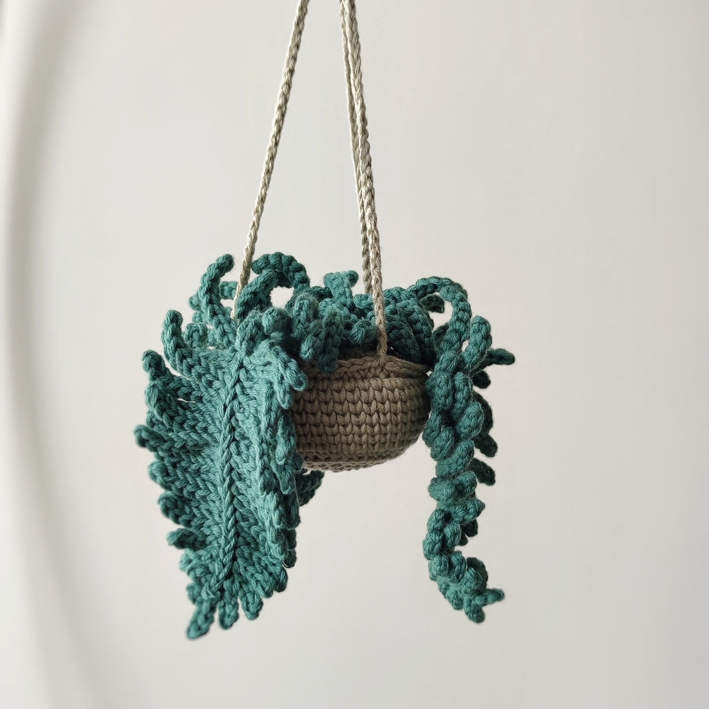 Crochet fern hanging plant pattern with textured green fern leaves and hanging cords on a neutral background.