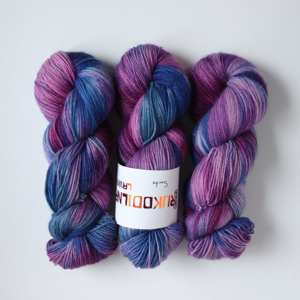 Hand dyed sock yarn in purple, violet, and deep blue tones with a knitted sock sample on wooden sock blocker, fingering weight 4-ply yarn
