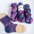 Hand dyed sock yarn in purple, violet, and deep blue tones with a knitted sock sample on wooden sock blocker, fingering weight 4-ply yarn