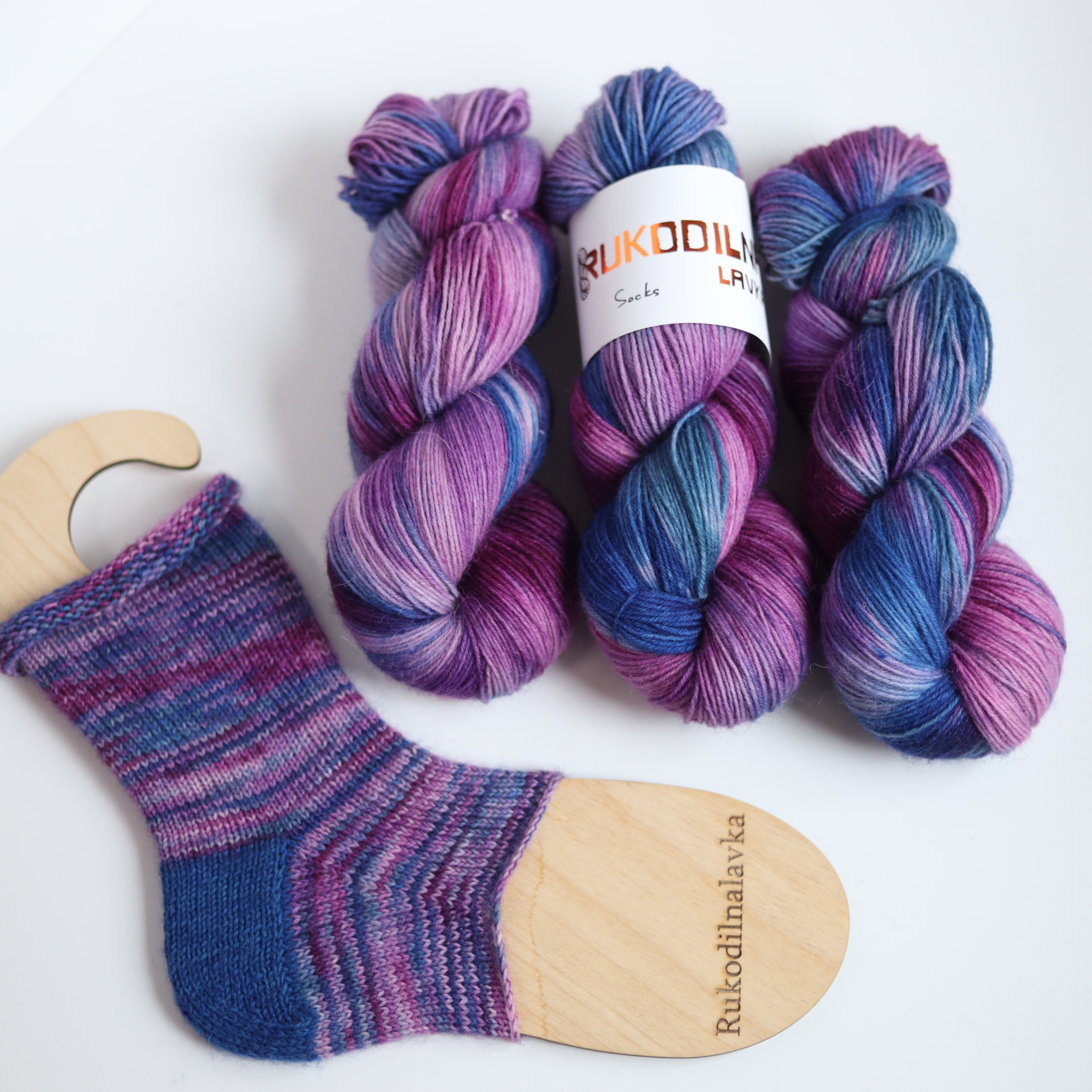 Hand dyed sock yarn in purple, violet, and deep blue tones with a knitted sock sample on wooden sock blocker, fingering weight 4-ply yarn