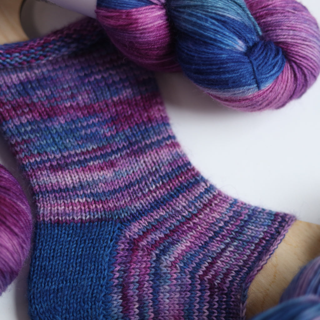 Hand dyed sock yarn in purple, violet, and deep blue tones with a knitted sock sample on wooden sock blocker, fingering weight 4-ply yarn