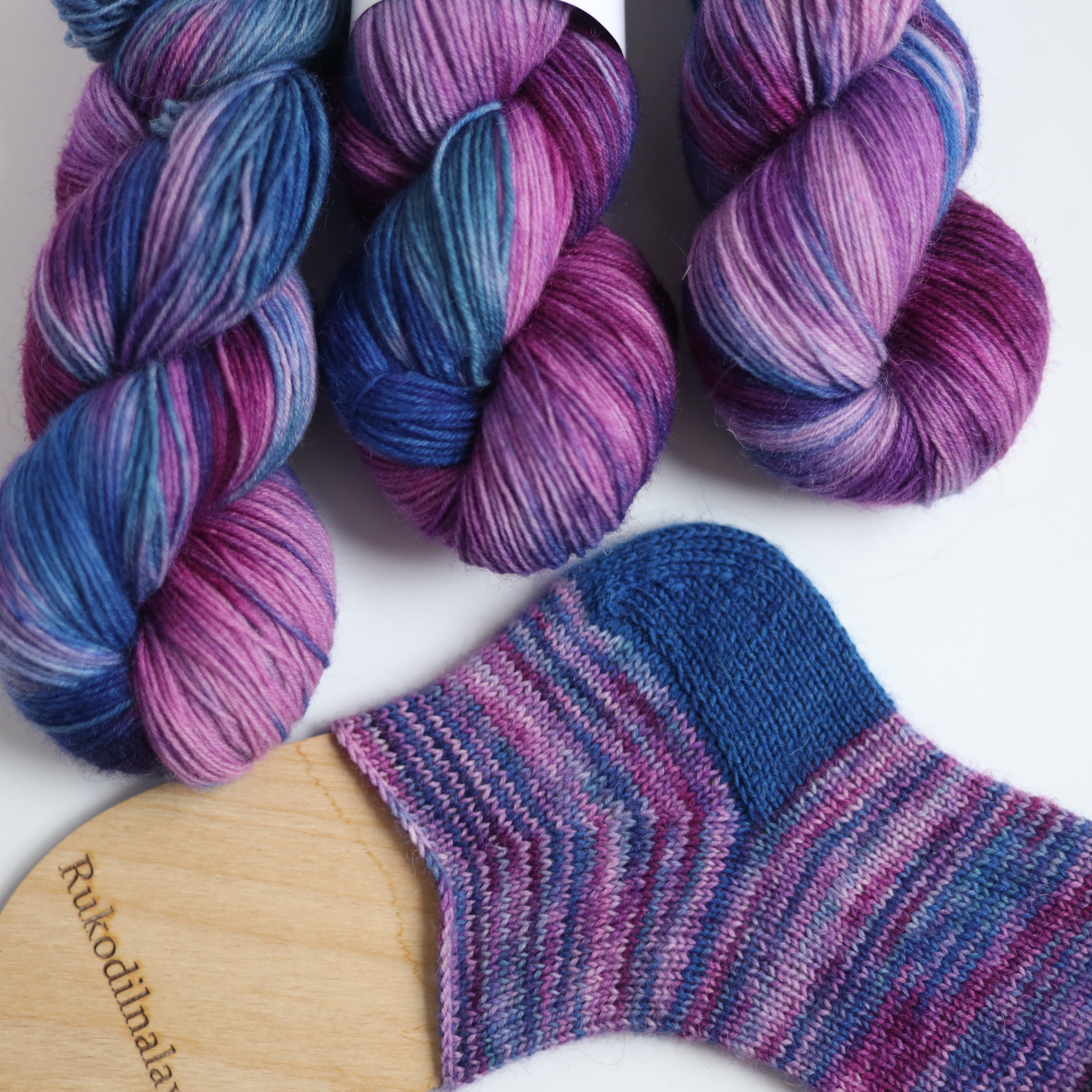 Hand dyed sock yarn in purple, violet, and deep blue tones with a knitted sock sample on wooden sock blocker, fingering weight 4-ply yarn