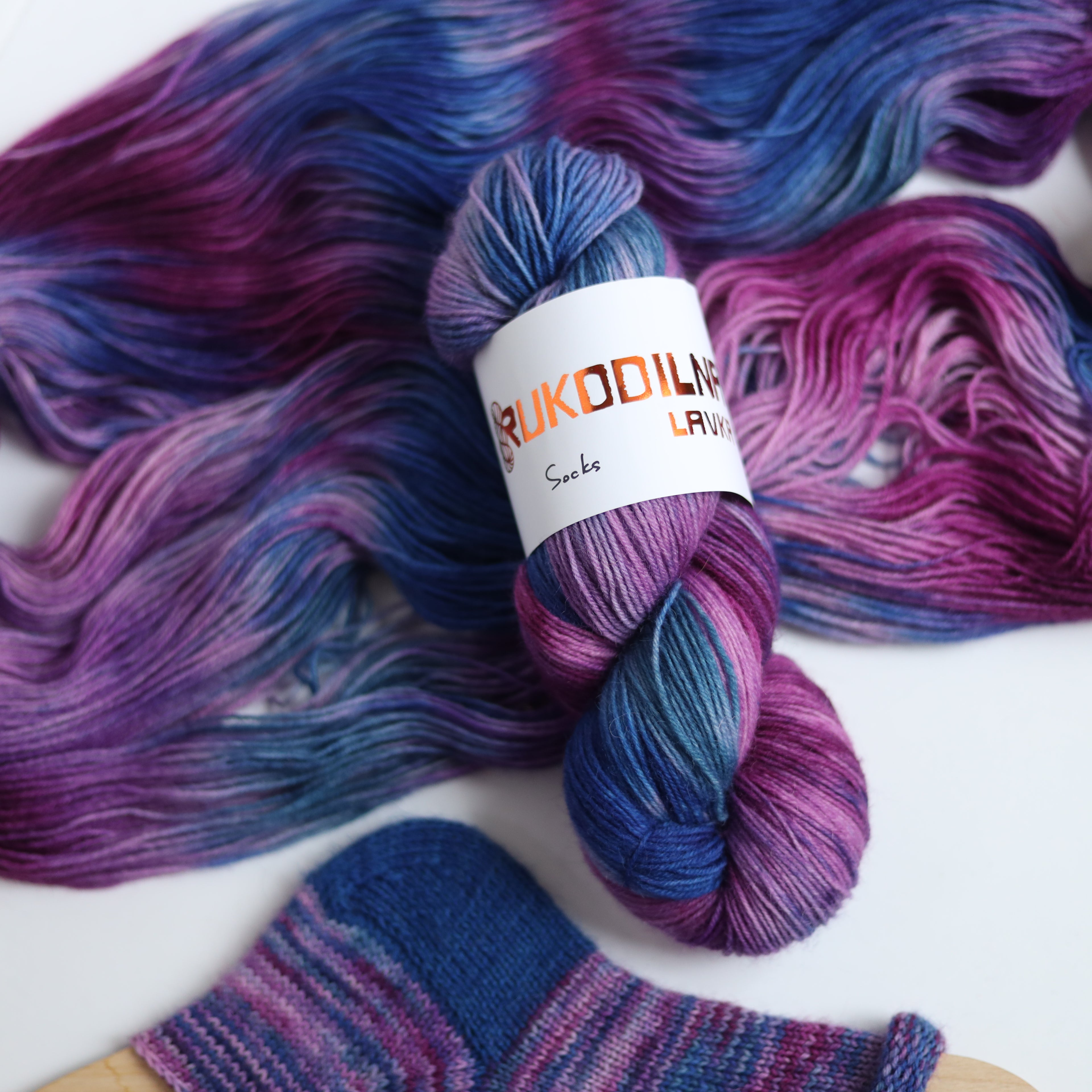 Hand dyed sock yarn in purple, violet, and deep blue tones with a knitted sock sample on wooden sock blocker, fingering weight 4-ply yarn