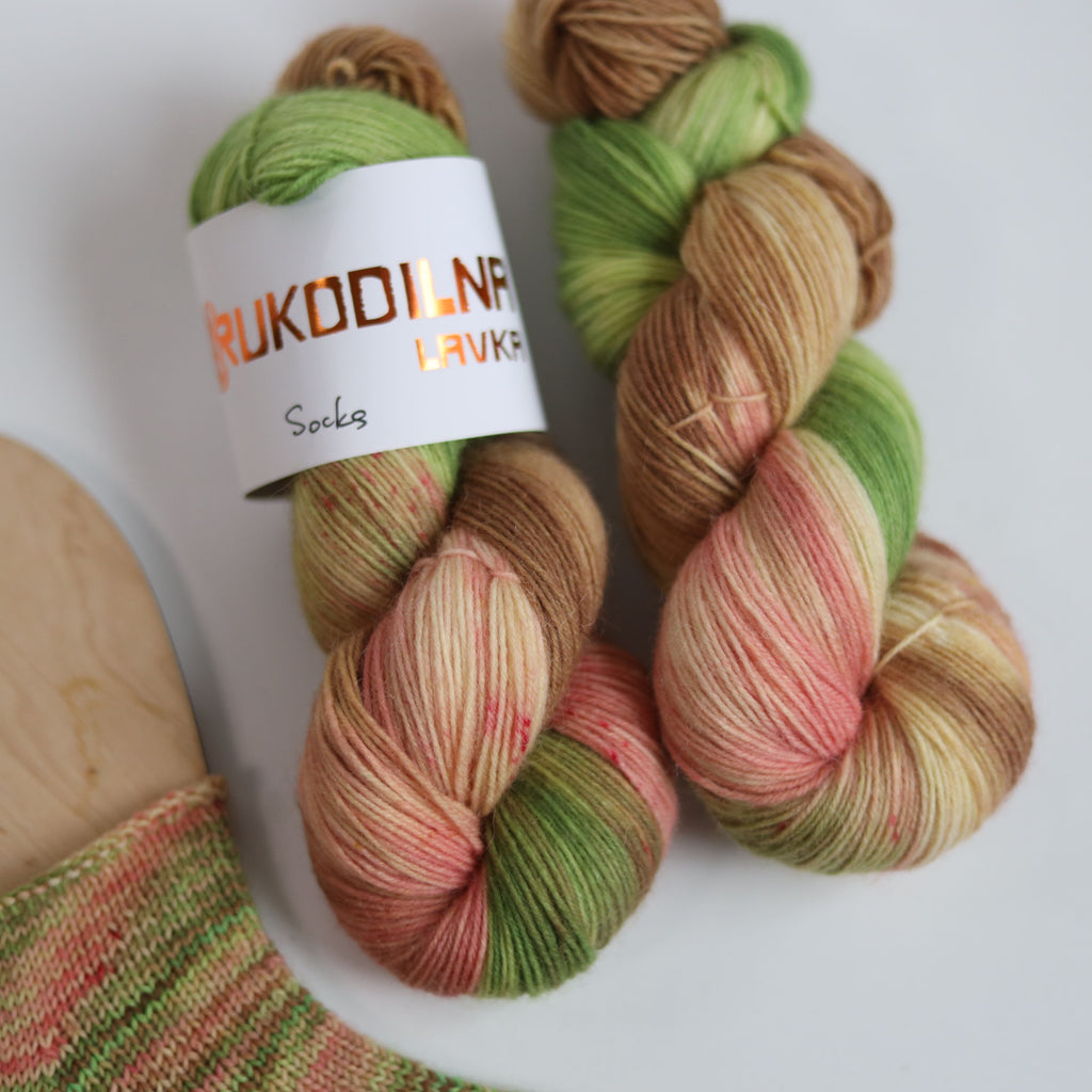 Hand dyed sock yarn in green, brown, and peach tones made from wool and polyamide, two skeins next to a knitted sock on a white background