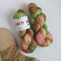 Hand dyed sock yarn in green, brown, and peach tones made from wool and polyamide, two skeins next to a knitted sock on a white background