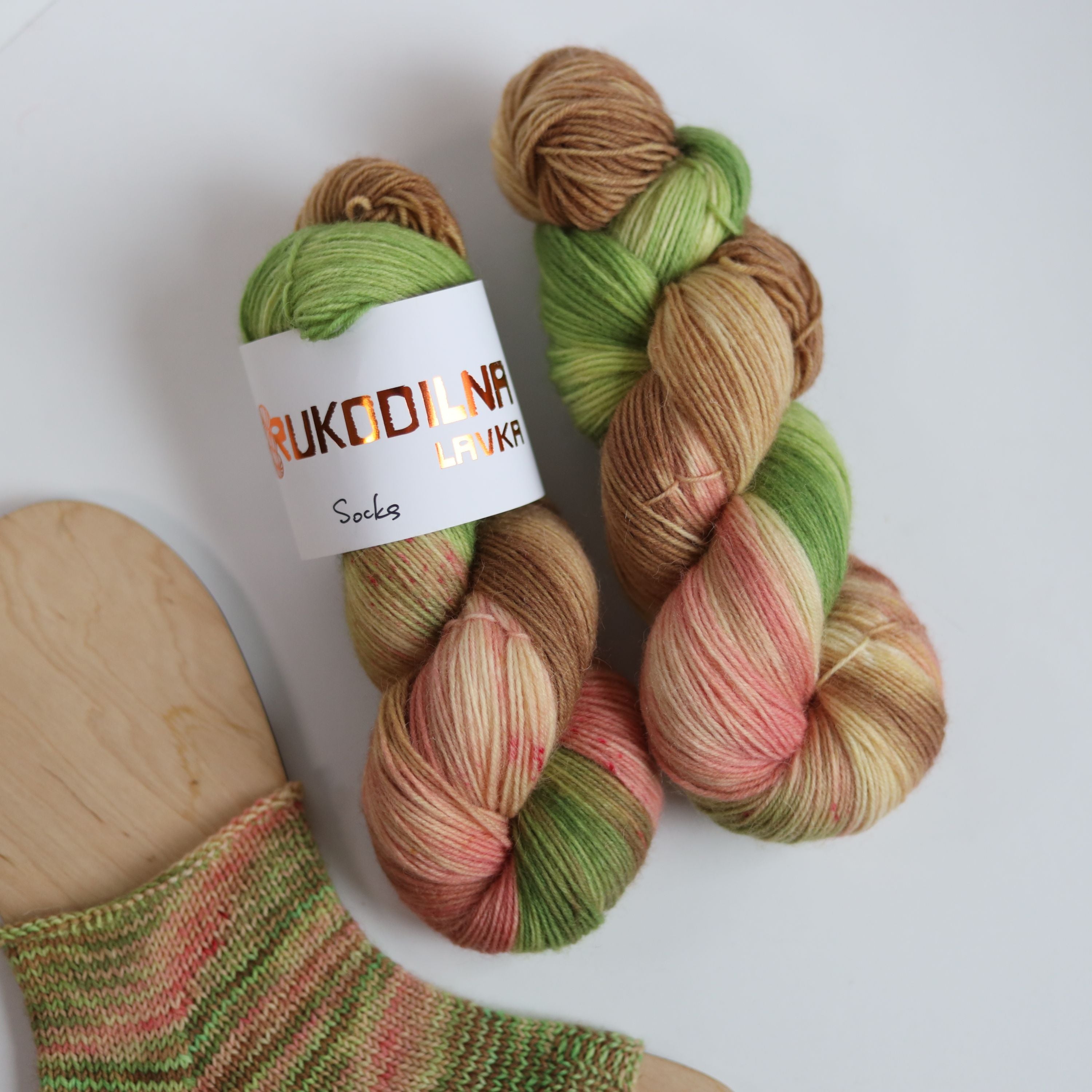 Hand dyed sock yarn in green, brown, and peach tones made from wool and polyamide, two skeins next to a knitted sock on a white background