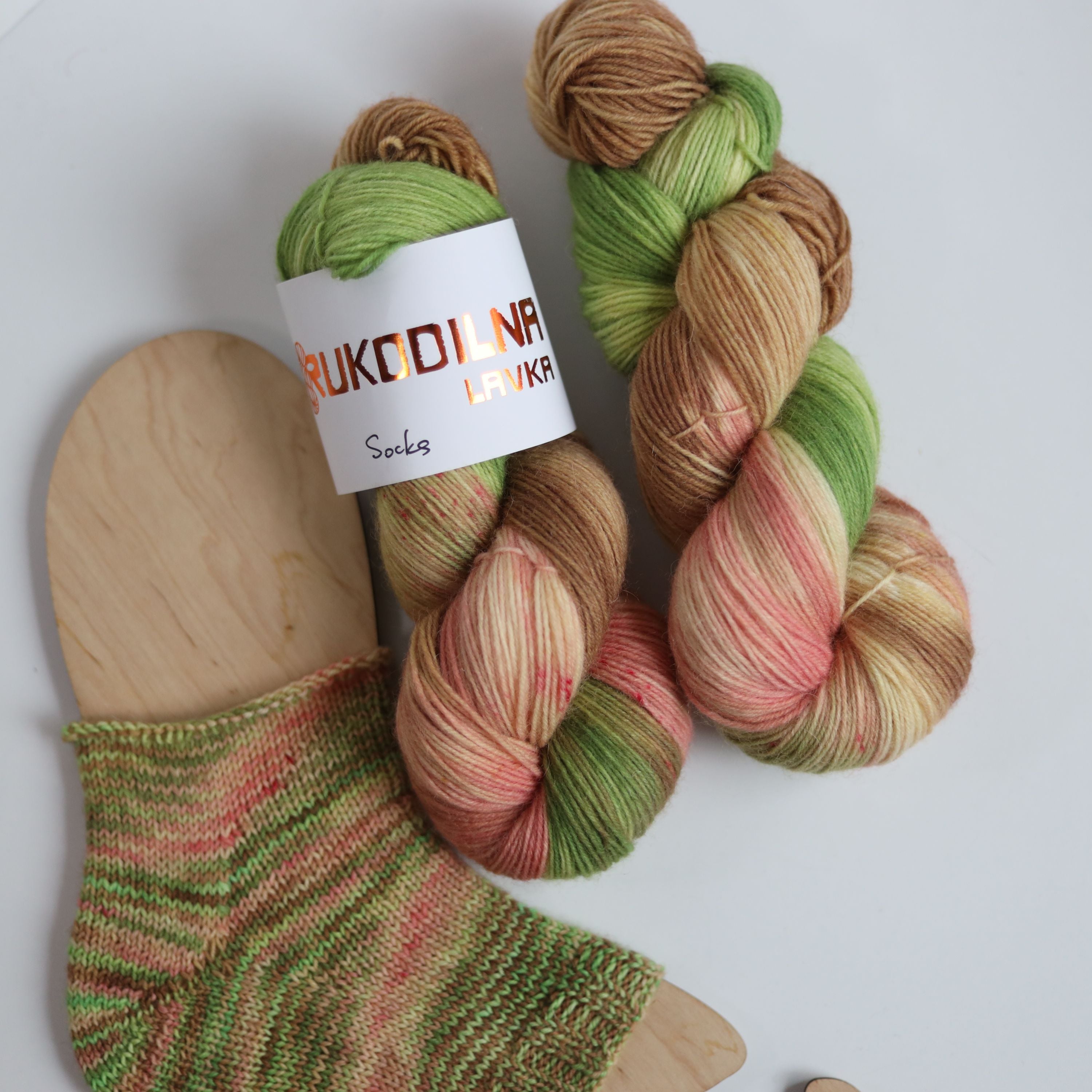 Hand dyed sock yarn in green, brown, and peach tones made from wool and polyamide, two skeins next to a knitted sock on a white background