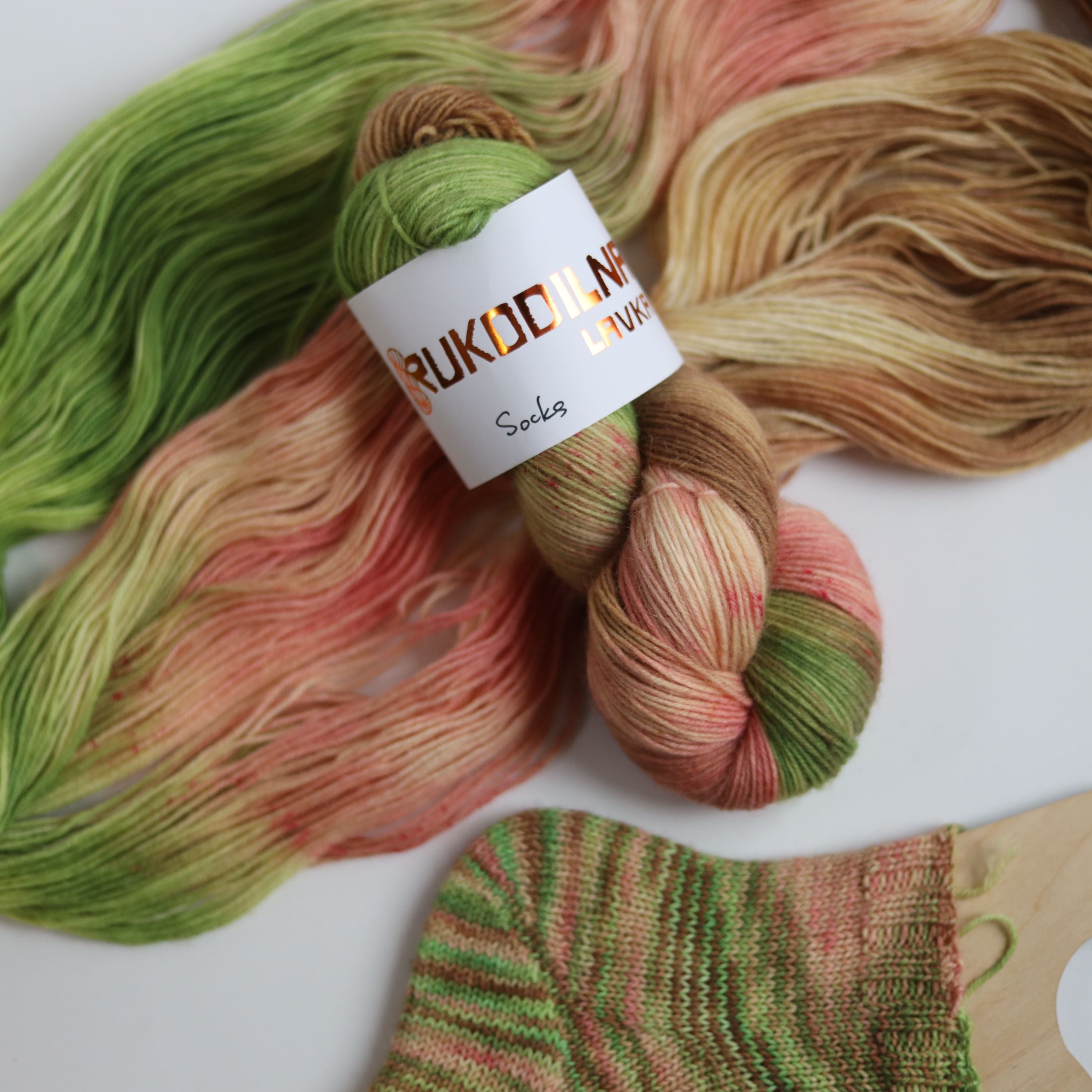 Hand dyed sock yarn in green, brown, and peach tones made from wool and polyamide, two skeins next to a knitted sock on a white background