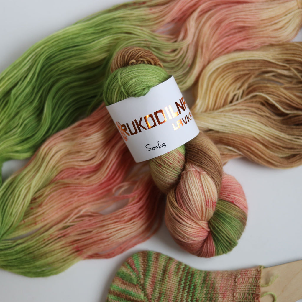Hand dyed sock yarn in green, brown, and peach tones made from wool and polyamide, two skeins next to a knitted sock on a white background