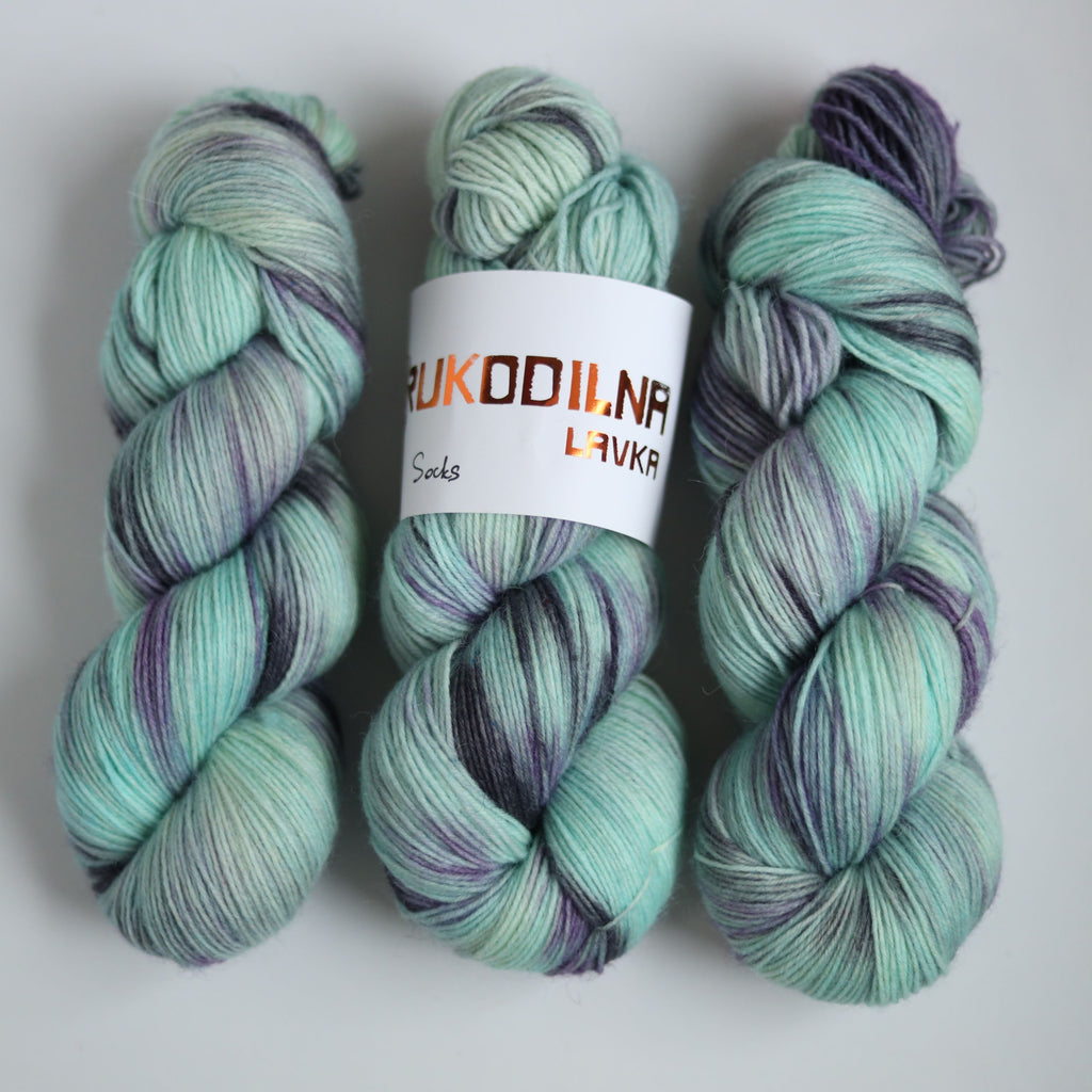 Three skeins of hand dyed green and purple variegated sock yarn made from superwash merino wool, shown with a knitted fabric sample on a white background.