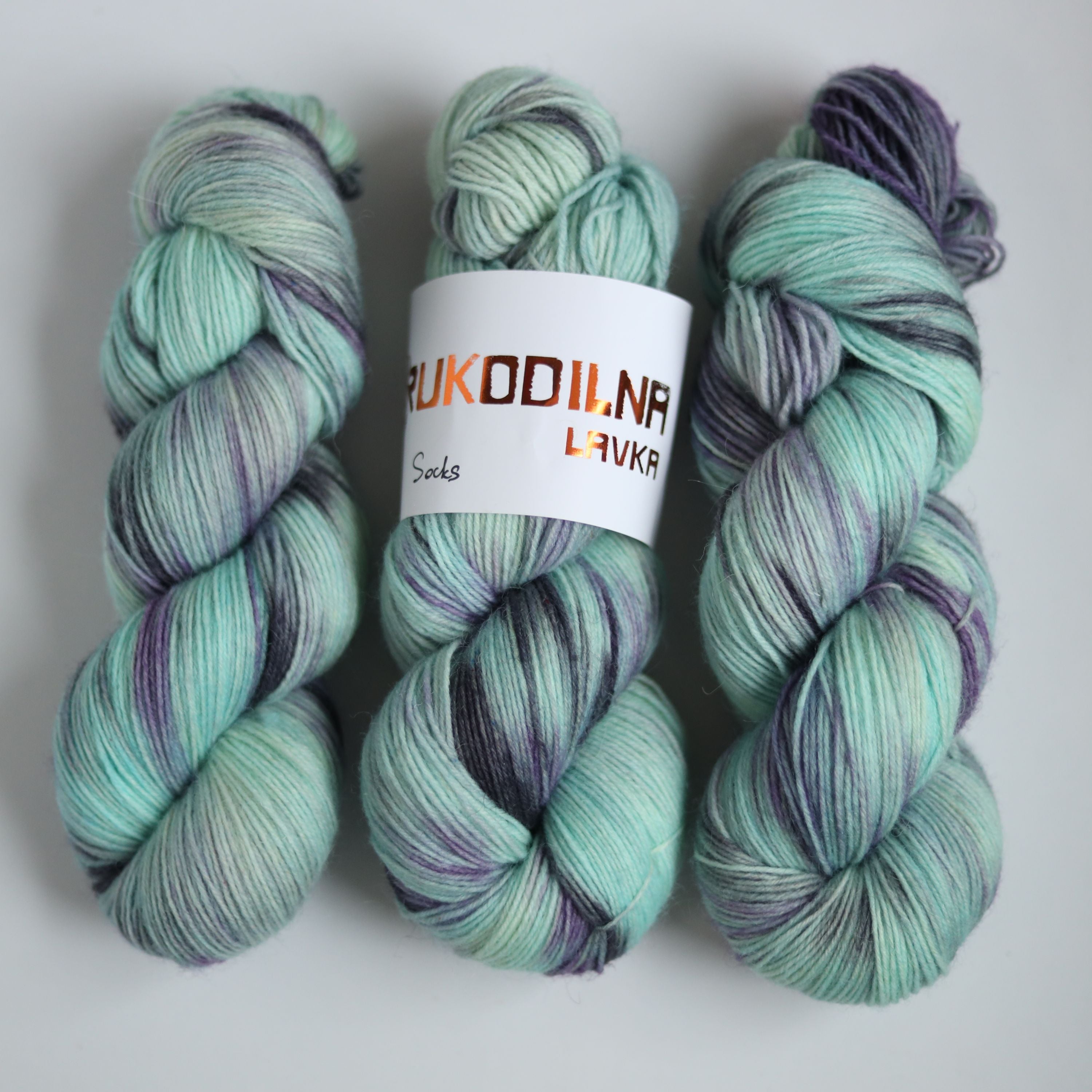 Three skeins of hand dyed green and purple variegated sock yarn made from superwash merino wool, shown with a knitted fabric sample on a white background.
