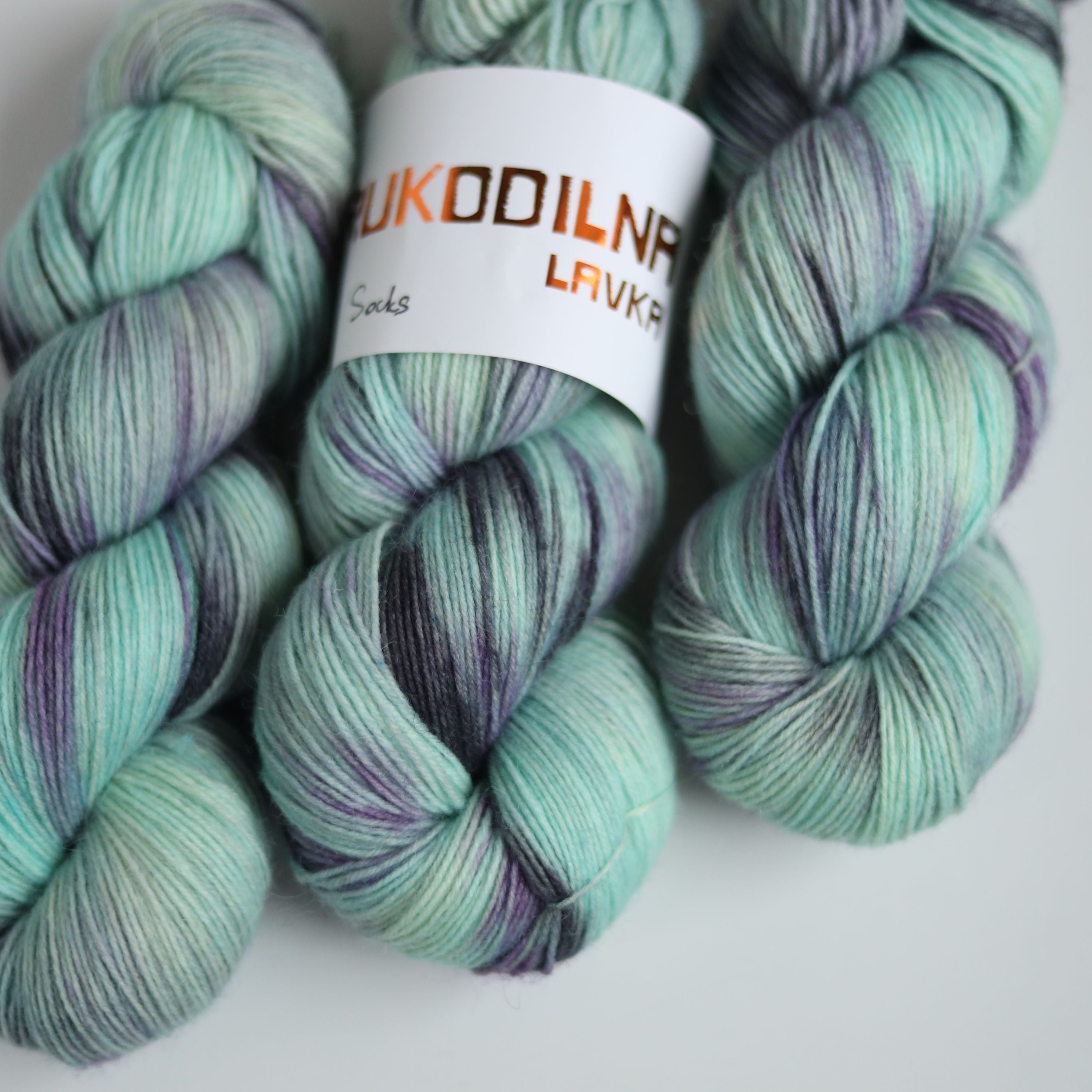 Three skeins of hand dyed green and purple variegated sock yarn made from superwash merino wool, shown with a knitted fabric sample on a white background.