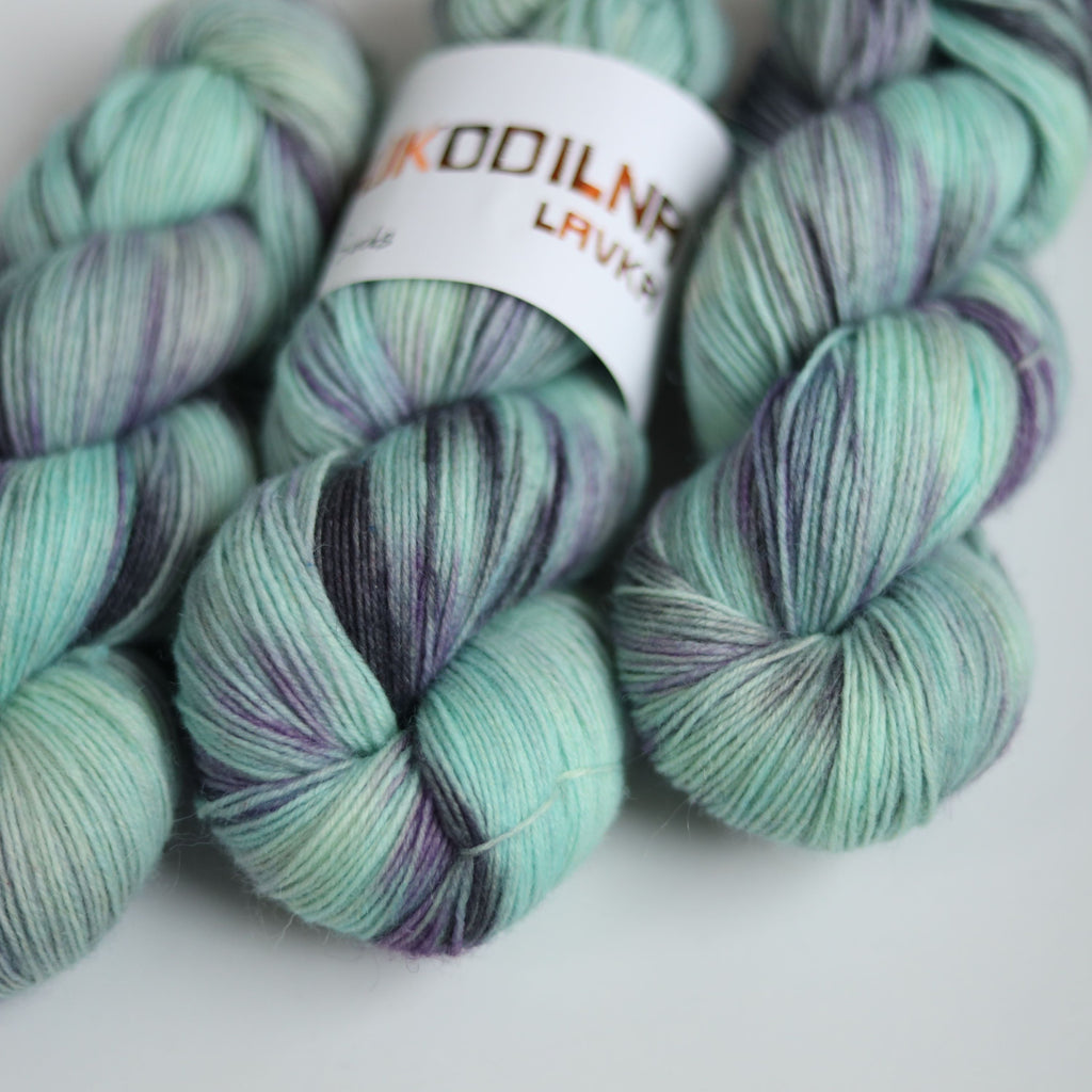 Three skeins of hand dyed green and purple variegated sock yarn made from superwash merino wool, shown with a knitted fabric sample on a white background.