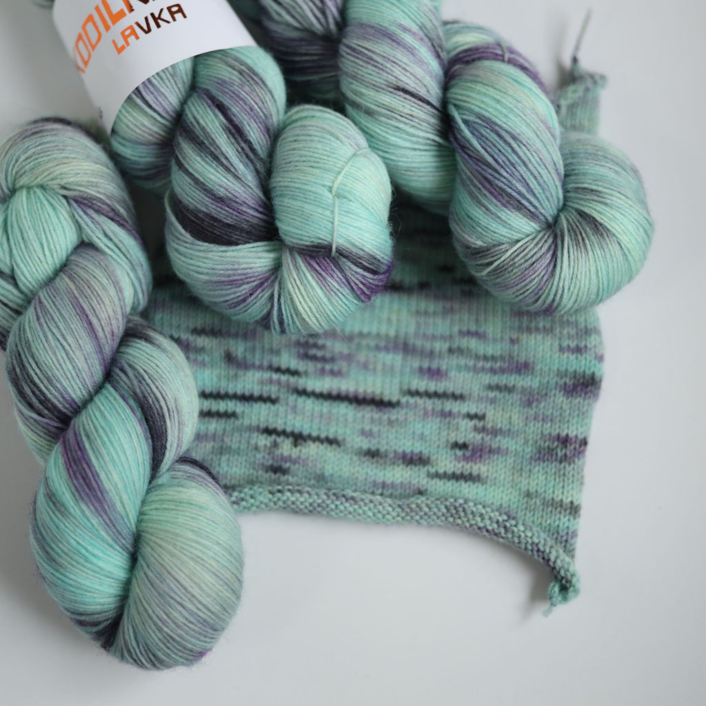 Three skeins of hand dyed green and purple variegated sock yarn made from superwash merino wool, shown with a knitted fabric sample on a white background.