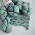 Three skeins of hand dyed green and purple variegated sock yarn made from superwash merino wool, shown with a knitted fabric sample on a white background.