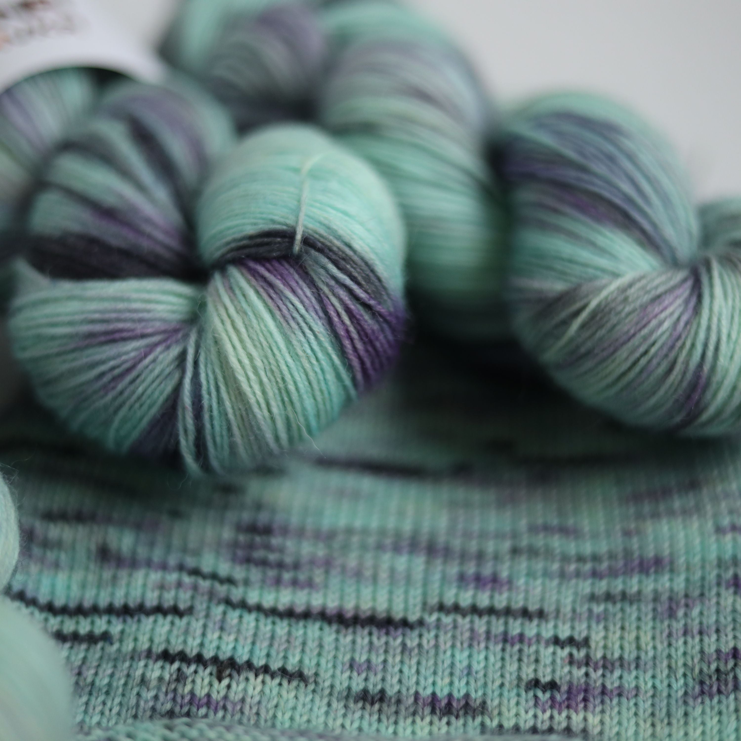 Three skeins of hand dyed green and purple variegated sock yarn made from superwash merino wool, shown with a knitted fabric sample on a white background.
