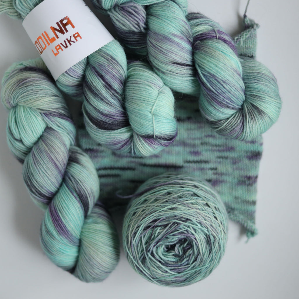 Three skeins of hand dyed green and purple variegated sock yarn made from superwash merino wool, shown with a knitted fabric sample on a white background.