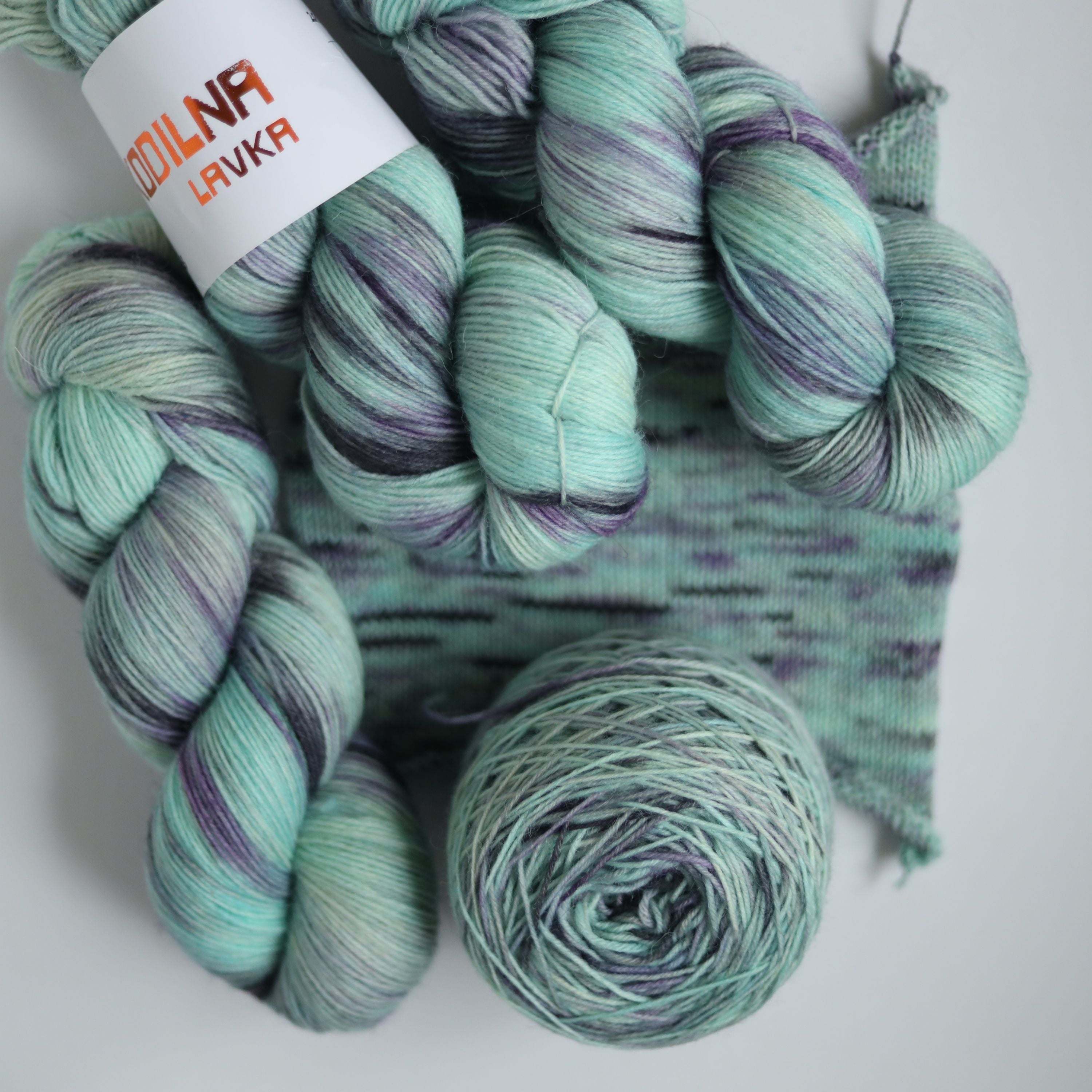 Three skeins of hand dyed green and purple variegated sock yarn made from superwash merino wool, shown with a knitted fabric sample on a white background.