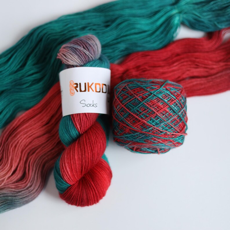 Hand dyed sock yarn in emerald green, red and lavender shades, fingering weight wool nylon yarn with knitted striped sample showing color transitions.