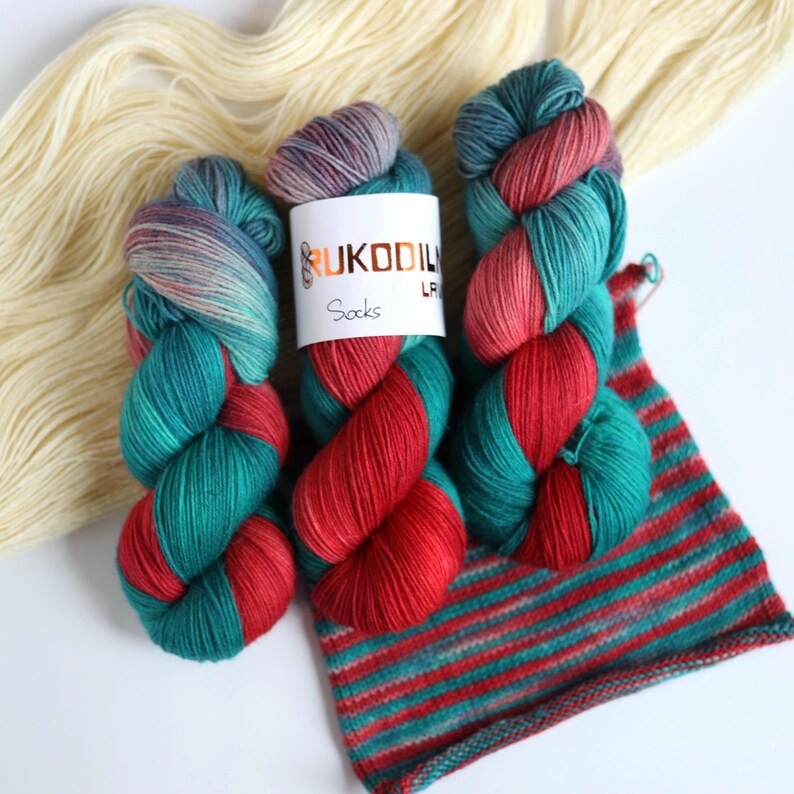 Hand dyed sock yarn in emerald green, red and lavender shades, fingering weight wool nylon yarn with knitted striped sample showing color transitions.