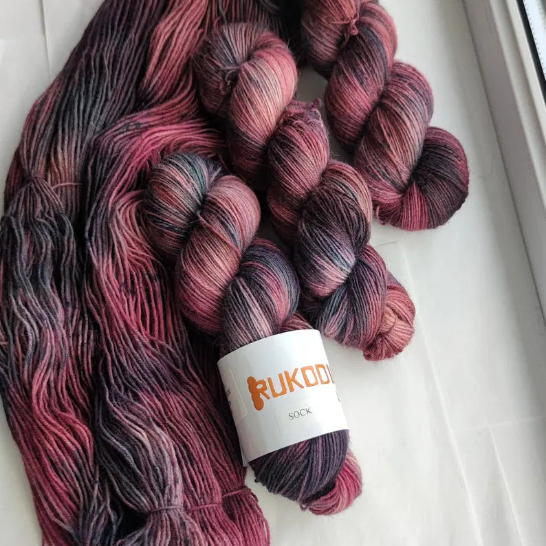 Hand-dyed sock yarn in blackberry, dusty rose and charcoal shades displayed as a variegated fingering weight wool nylon skein.